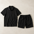 a coordinated black men's two-piece outfit: one plain short-sleeve polo shirt with classic collar and button placket AND one pair of matching black drawstring shorts, neatly arranged side by side, both fully visible