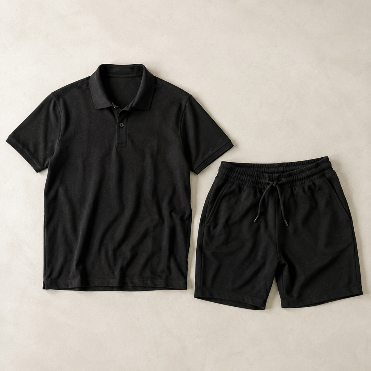 a coordinated black men's two-piece outfit: one plain short-sleeve polo shirt with classic collar and button placket AND one pair of matching black drawstring shorts, neatly arranged side by side, both fully visible