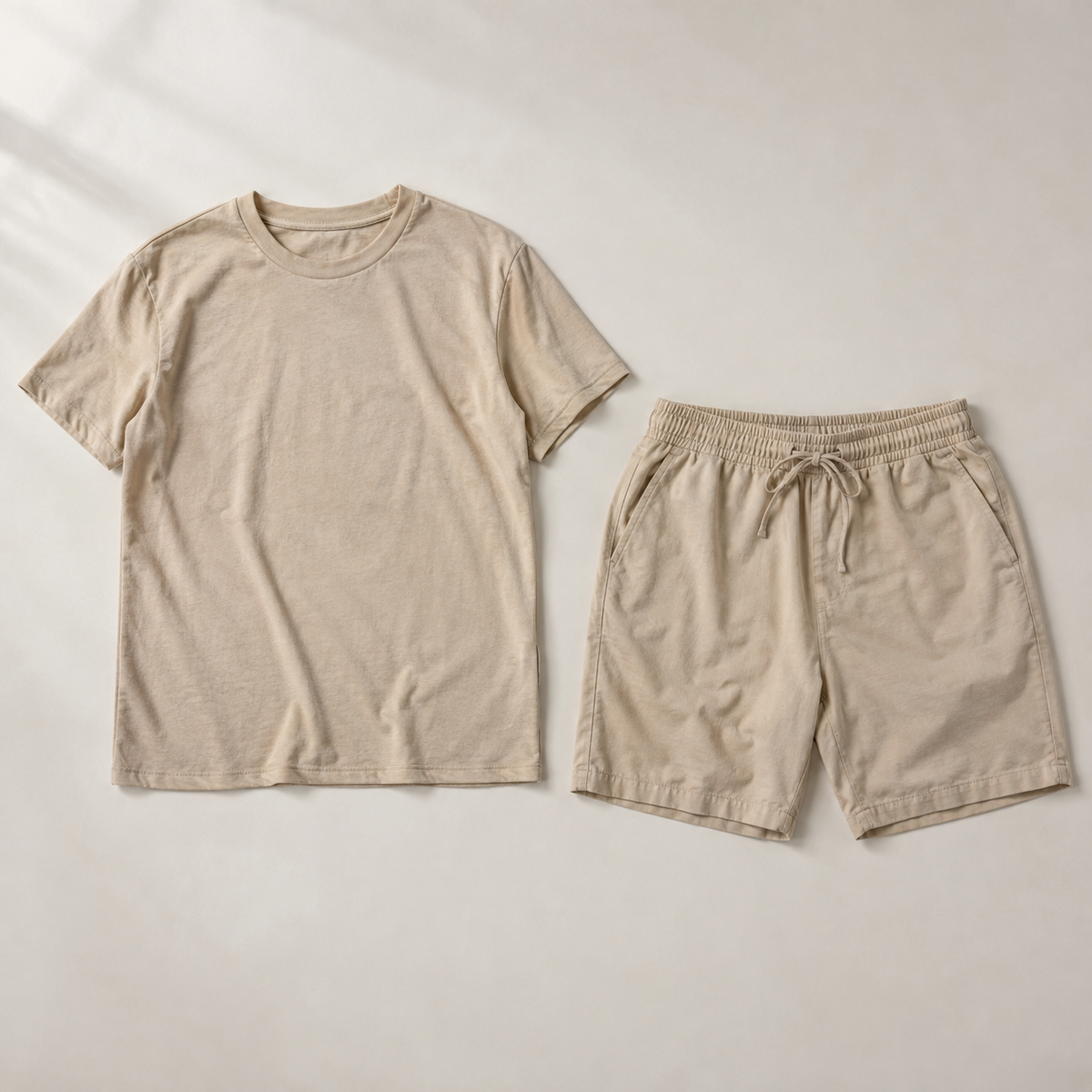 a coordinated beige men's two-piece outfit: one plain crew-neck short-sleeve t-shirt AND one pair of matching beige drawstring shorts, neatly arranged side by side, both fully visible